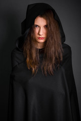 Fototapeta premium Young, beautiful girl in a black cloak with a hood in the studio on a gray background. Long hair fluttering on the wind. A model with clean skin. Suit of a fortuneteller, witches for Halloween.