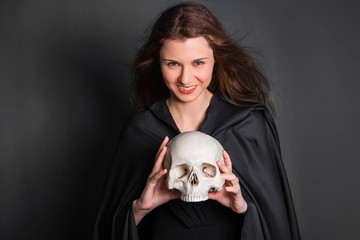 Beautiful, young girl with long hair in a black cloak with a skull in her hands. Studio photo on a gray background. Witchcraft, necromancy, fortune telling, Halloween.