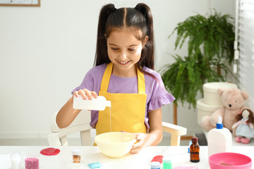 Cute little girl pouring glue into bowl at table in room. DIY slime toy
