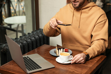 Male blogger taking photo of dessert and coffee at table in cafe, closeup