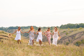 Obraz premium The company of cheerful female friends have a great time together on a picnic in a picturesque place overlooking the green hills. Girls in white dresses dancing in the field
