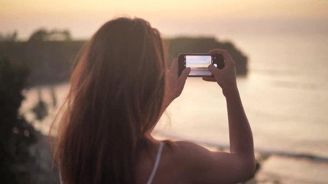 Close up rear view of young woman making photos and videos of beautiful ocean sunset. Attractive female takes pictures standing on seashore at sundown. Woman taking pictures of beach at sunset 4k