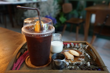 Close up plastic take away cup of iced black coffee served with milk, sugar syrup and butter cookies on wooden tray table decorated with dry flower and coffee beans