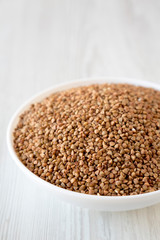 Uncooked Roasted Buckwheat in a white bowl on a white wooden background, side view. Copy space.