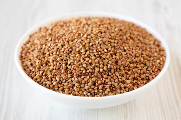 Uncooked Roasted Buckwheat in a white bowl on a white wooden surface, side view. Close-up.