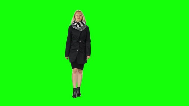 Blonde girl in a black dress, coat, scarf and boots going against a green screen.