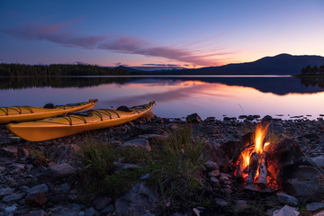 Campfire during dusk at the shore of a lake with two kayaks. Sweden.