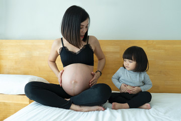 Mother and daughter sit on bed showing  tummy.