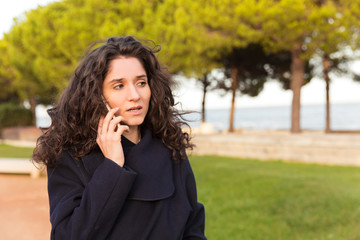 Thoughtful woman talking on smartphone while walking on street. Serious curly lady wearing coat communicating via phone. Technology, communication concept