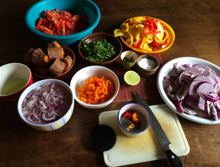 Bowls and vegetables on a brown table including chopped chilies, onions, peppers, tomatoes, sweet potatoes and lemon in different warm colors