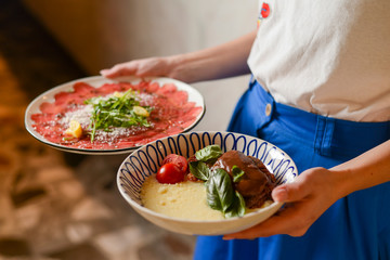 Carpaccio of beef served on a white plate. Waiter holds two plates with Italian food in Italian restaurant.
