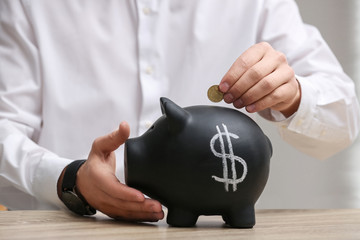 Man putting coin into piggy bank with dollar sign at wooden table, closeup