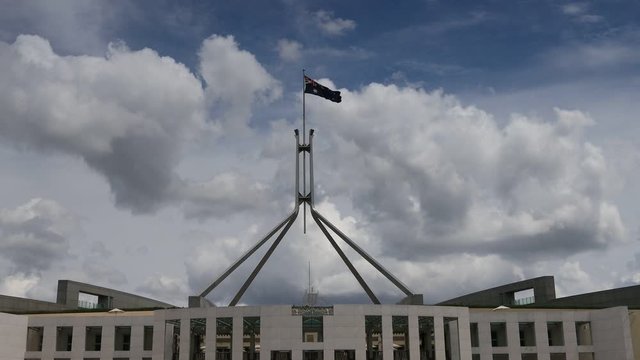 Australian national flag on tall flagpole agains blue sky waving over the federal parliament house.