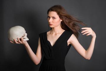 A beautiful, young girl with long hair in a black dress with a skull in her hands. Studio photo on a gray background. Witchcraft, necromancy, fortune telling, Halloween.