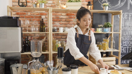 Smiling female barista putting espresso on tray in counter in bar. young girl waitress preapre customer order in morning coffee shop. woman staff in apron make cup of latte beside tasty croissant