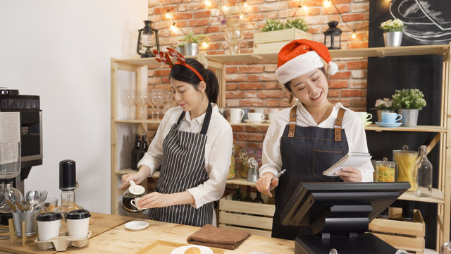 happy two asian waitress wearing santa claus hat celebrating christmas event. young girl barista in cute reindeer horns on head making coffee pour into cup. cafe bar staffs working on xmas holidays