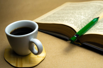 White cup with coffee on a wooden stand with a book and a green ballpoint pen on a beige background