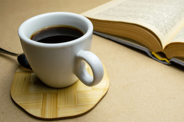 White cup with coffee on a wooden stand with a spoon with a book on a beige background