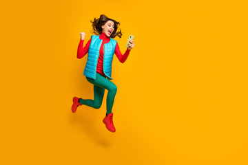 Fototapeta na wymiar Full size photo delighted girl use smartphone follow social media autumn discount win lottery raise fists scream jump run wear green blue red pants shoes vest isolated yellow color background