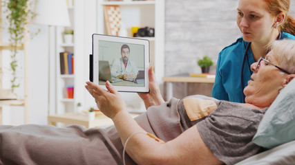Old disabled woman lying in hospital bed having an online video call with a doctor