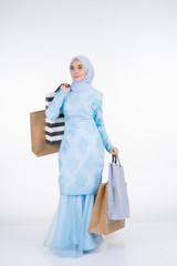 A beautiful Muslim female model in a Asian traditional dress modern kurung carrying shopping bags isolated on white background. Eidul fitri festive preparation shopping concept. Full length portrait.