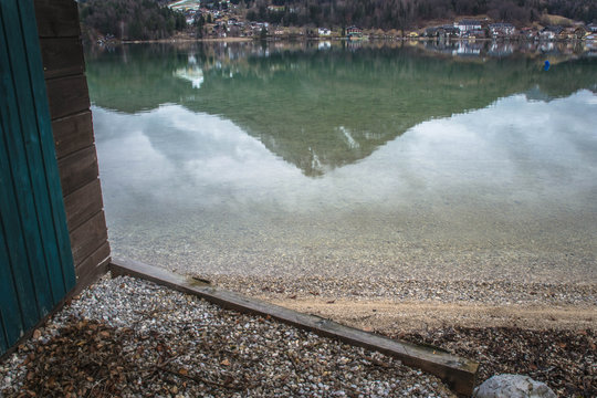 Soft lake reflections of mountains and a lakeside village viewed next to the corner of a wooden boathouse. Quiet, moody waterside atmosphere.