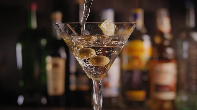 Close-up of a Martini glass with olives and lime poured Martini or gin on the bar on a blurred background of bottles of various alcohol. Slow motion. The glass turns slowly.