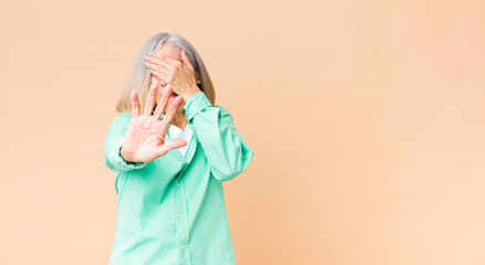 middle age cool woman covering face with hand and putting other hand up front to stop camera,...