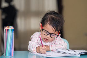 little girl wearing glasses drawing on book at table.