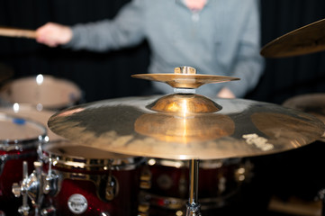 Shiny gold-colored cymbal and shimmering red drums with a man in green vest playing drums behind it.