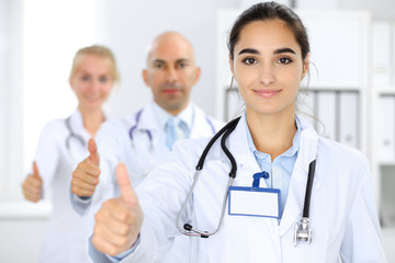 Fototapeta premium Doctor woman showing OK in clinic with colleagues at the background. Hispanic or latin american staff in medicine