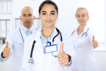 Doctor woman showing OK in clinic with colleagues at the background. Hispanic or latin american staff in medicine