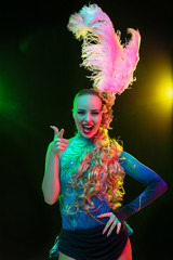 Pointing. Beautiful young woman in carnival, stylish masquerade costume with feathers on black background in neon light. Copyspace for ad. Holidays celebration, dancing, fashion. Festive time, party.