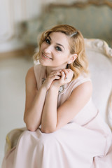 Fototapeta premium Beautiful blonde woman in a delicate pink dress sits on a chair in a bright room.