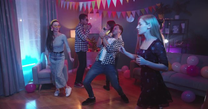 Team of young stylish and happy guys and girls dancing and having fun while having fun at home party. Guy imitating singing in the mic. Indoors.