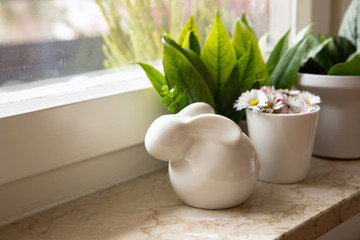 White porcelain easter bunny lay on the windowsill with green leaves and blooming daisies in flowerpot. Easter home decoration.