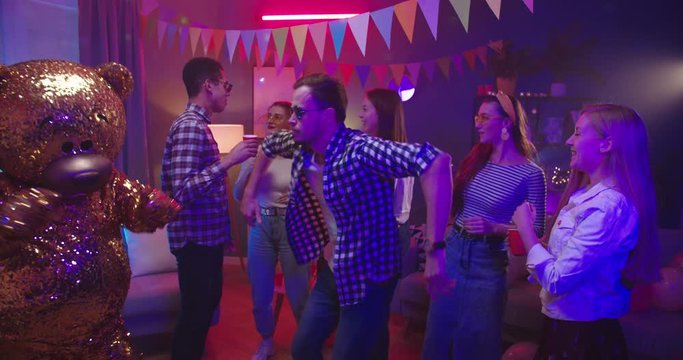 Party at home: cheerful young Caucasian guy dancing with a big golden grown doll of teddy bear and having fun at the apartment while resting with friends.