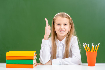 Smart schoolgirl raising hand during on the background of a school board