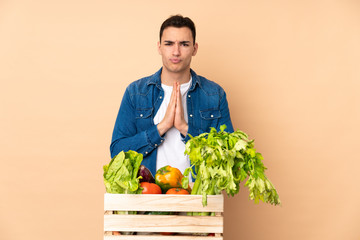 Farmer with freshly picked vegetables in a box isolated on beige background keeps palm together. Person asks for something