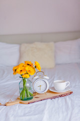 A bouquet of gerberas in a glass jar, a cup of cappuccino, a white alarm clock on a wooden stand stand on the bed. Breakfast in bed.
