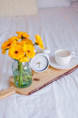 A bouquet of gerberas in a glass jar, a cup of cappuccino, a white alarm clock on a wooden stand stand on the bed. Breakfast in bed.