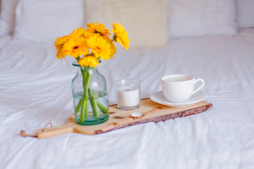 A bouquet of gerberas in a glass jar, a cup of cappuccino, a clock, a candle on a wooden stand are on the bed. Breakfast in bed.