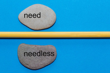 The words need and needless are written on natural smooth stones separated by a yellow pencil. The...