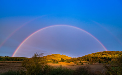 Fototapeta na wymiar Rainbow after rain hanging in the blue sky; beautiful scenery in the field