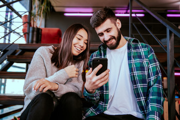 On the modern work office studio one good looking guy and pretty lady smiling have a break time sitting on the stairs have, watching a funny video