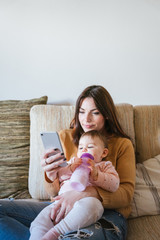 Portrait of a mom and her baby on the sofa - The mother and daughter look at the smart phone, the...