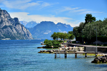 Malcesine, Italia - Lake Garda, on the right is a promenade with green trees, a pier, in the background alpine mountains, a blue sky with white clouds, in the summer afternoon.