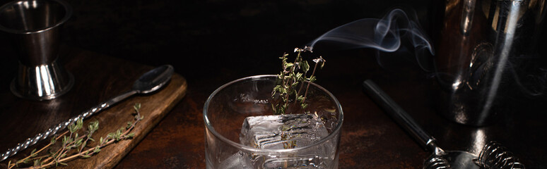 transparent glass with burning herb with smoke and ice cube on wooden surface, panoramic shot