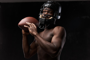 Fototapeta na wymiar portrait photo of dark-skinned young man with nude torso on a dark background he has a rugby helmet on head and holds a rugby ball up in his arm