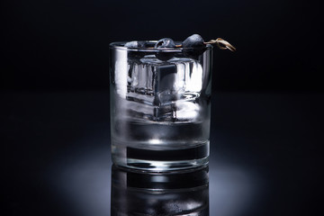 transparent glass with ice cube and vodka garnished with blueberries on black background